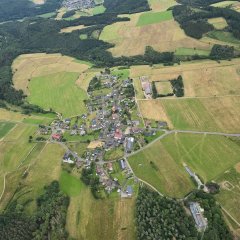 Ein Bild aus der Vogelperspektive zeigt den Ort Winnerath im Sommer. Umgeben von grünen Wiesen, Feldern und Wäldern strahlen die Häuser mit vereinzelten roten Dächern in der Sonne. Im Vordergrund erkennt man den Sportplatz. 