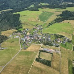 Ein Bild aus der Vogelperspektive zeigt den Ort Winnerath im Sommer. Umgeben von grünen Wiesen, Feldern und Wäldern strahlen die Häuser mit vereinzelten roten Dächern in der Sonne. Im Vordergrund erkennt man den Sportplatz. 